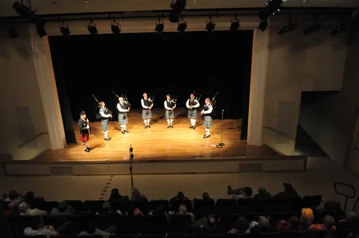 A pipe band performs on a brightly lit theater stage before a seated audience.