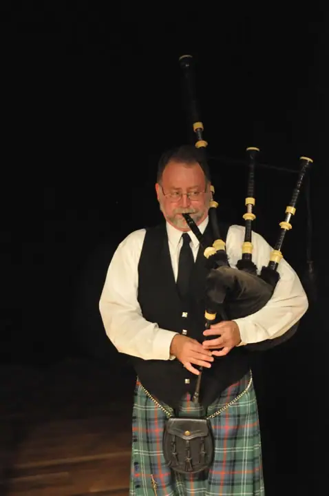 A man in a tartan kilt and vest plays bagpipes against a dark background.