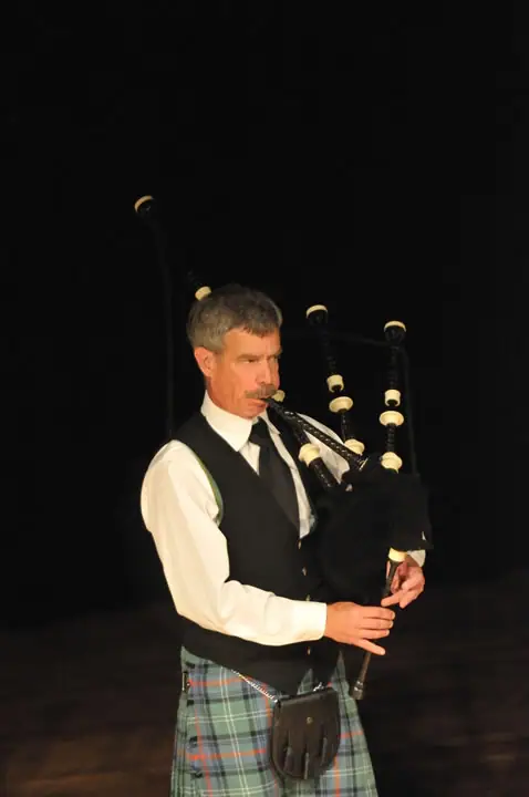 A musician in a kilt plays bagpipes against a dark background.