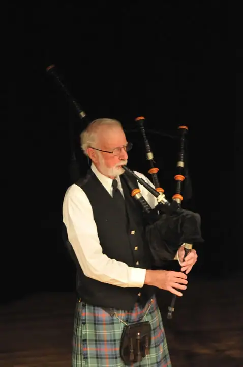 Bearded musician in glasses plays bagpipes while wearing a tartan kilt and black waistcoat.