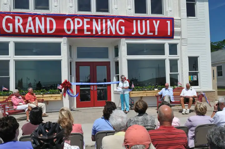 A speaker addresses a seated crowd outside a building decorated for a grand opening.
