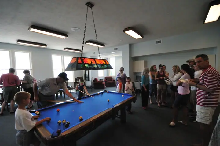 Adults and children gather around a blue-felt pool table in a bright room.