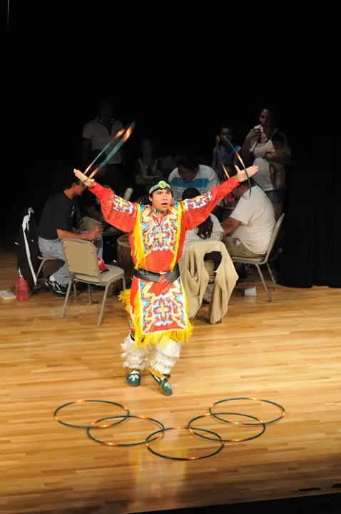 A performer in colorful regalia raises two hoops above a wooden stage floor surrounded by more hoops.