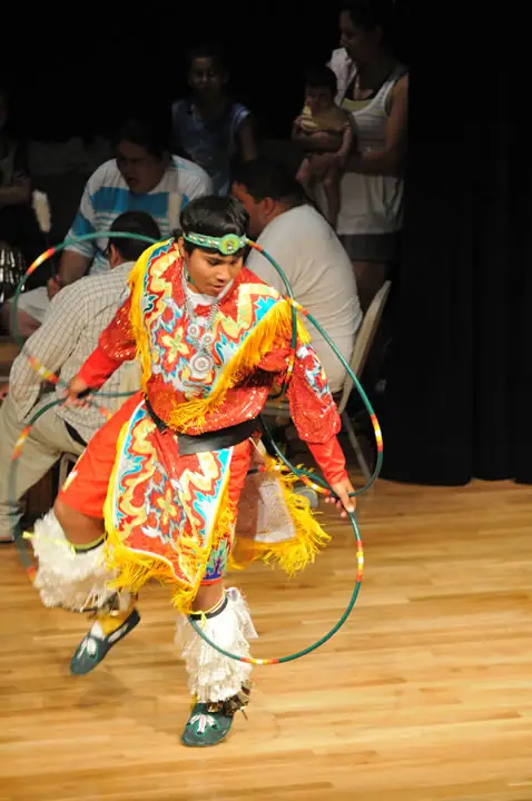 A young dancer in colorful fringed regalia performs with hoops before an audience.