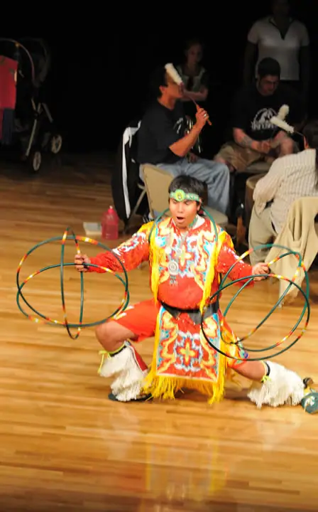 Young performer in a colorful fringed outfit kneels on a wooden floor holding multicolored hoops.