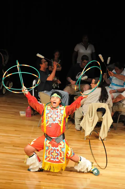 A costumed performer kneels on a wooden floor while holding two colorful hoops before seated onlookers.