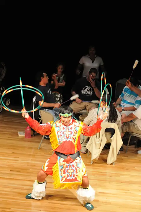 A child in colorful fringed attire performs with hoops before a seated audience indoors.