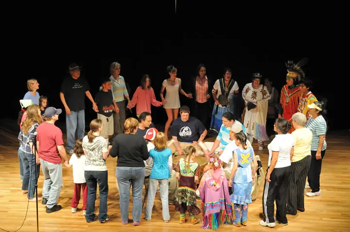Adults and children form a circle on a stage, with several people wearing colorful regalia and feathered headdresses.