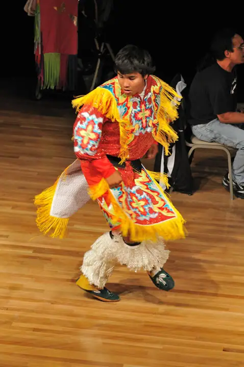 A dancer in a colorful fringed costume moves across a wooden floor while people sit nearby.