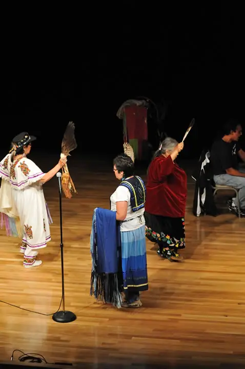 People in colorful garments hold feathered fans and fringed cloths on a wooden stage.