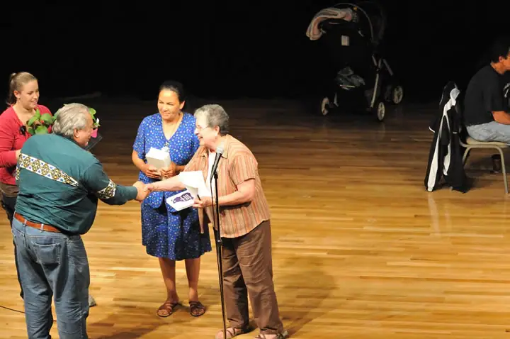 People exchange a handshake and papers on a polished wooden stage while others stand nearby.