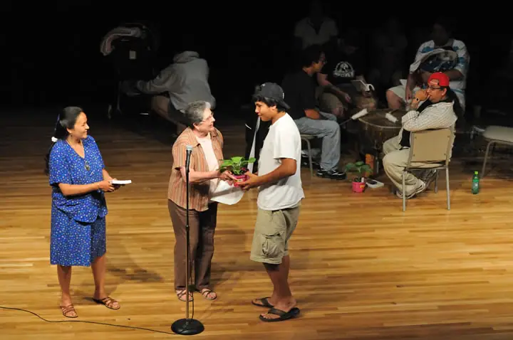 Adults gather on a stage as a young man receives a potted plant during a presentation.