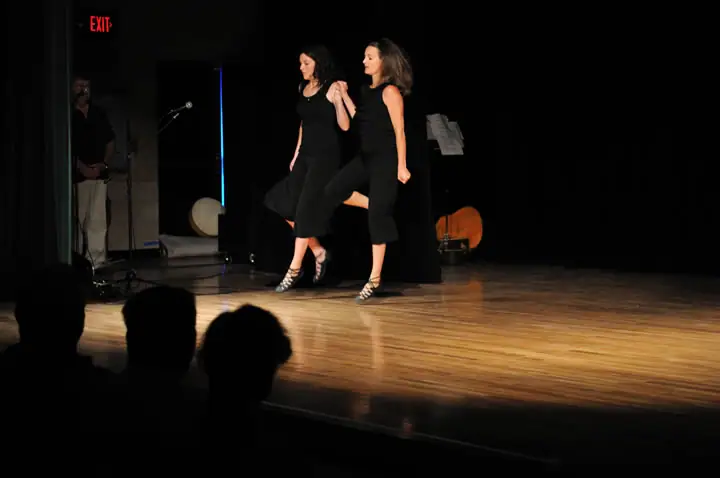 Two women perform a synchronized dance on a stage before a seated audience.