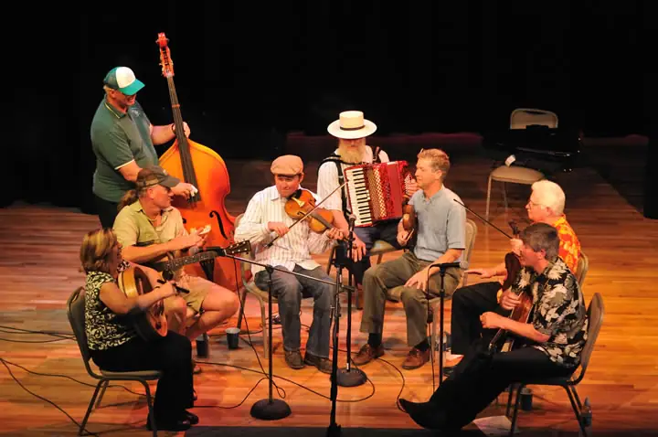 Eight musicians perform together on a wooden stage with guitars, violin, accordion, and double bass.