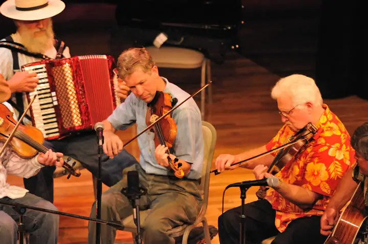 Musicians play accordion and violins on a wooden stage, with microphones and another string instrument visible.