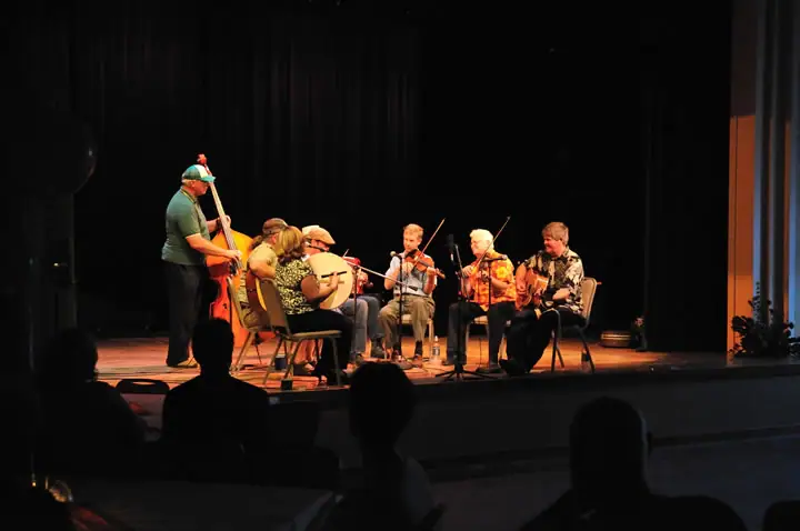 Musicians perform on a lit stage while audience members watch in silhouette.