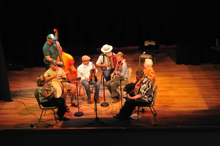 Musicians perform together on a warmly lit wooden stage with banjo, double bass, accordion, fiddles, and guitar.