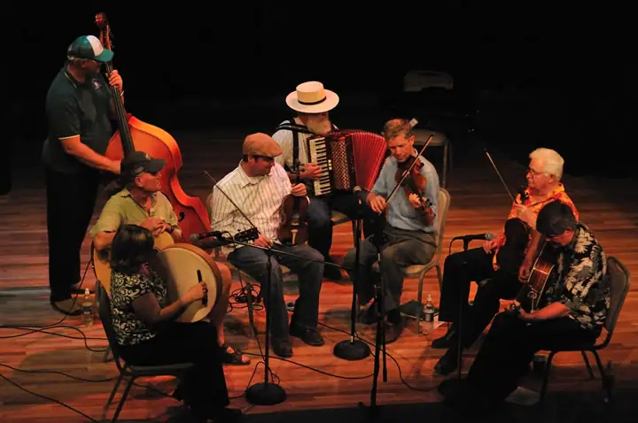 Musicians perform together on a wooden stage, playing strings, accordion, and percussion.