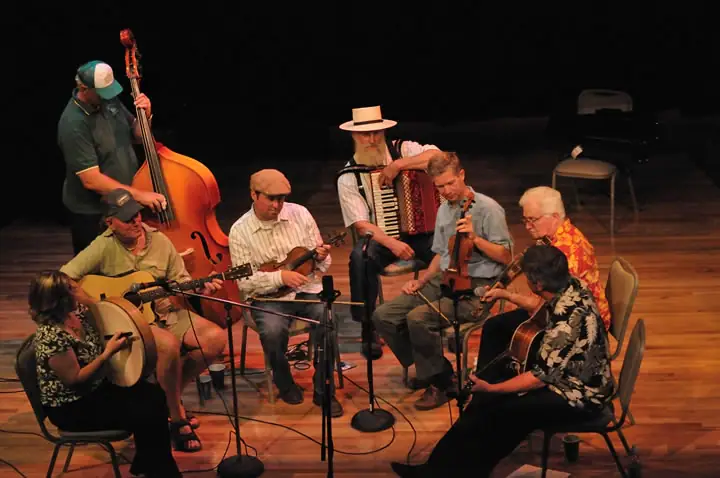 A group of musicians performs on a stage with string instruments, an accordion, microphones, and chairs.