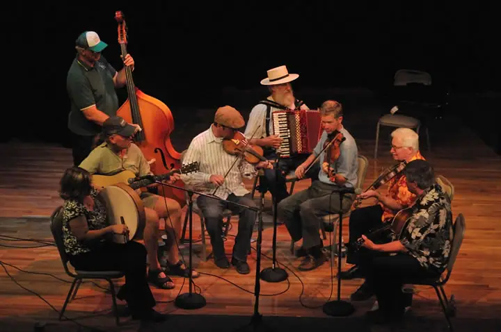 Musicians perform together on a stage with fiddles, guitars, an accordion, a double bass, and a frame drum.