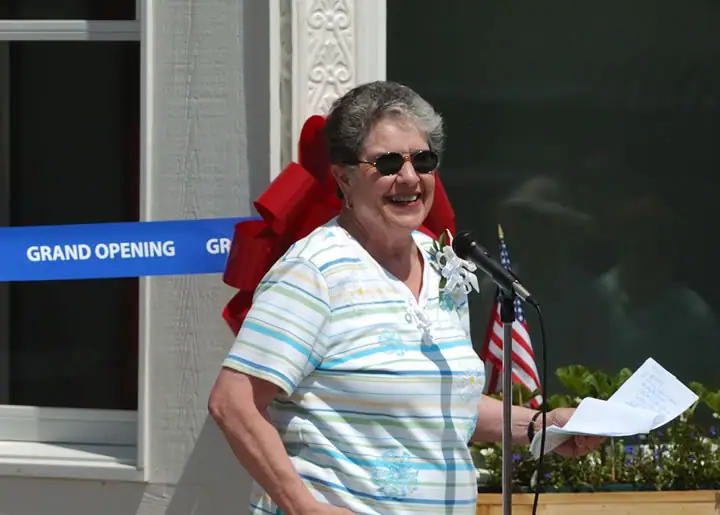 A woman speaks into a microphone beside a blue grand-opening ribbon and a large red bow.