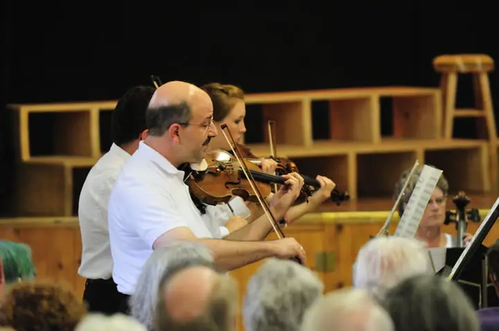 Musicians play violins before an audience in a wood-paneled performance space.