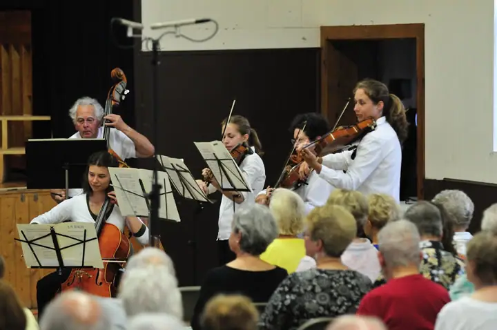 Musicians perform string music before a seated audience in an indoor hall.