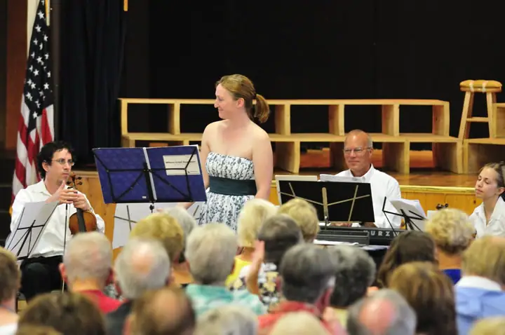 A woman and two seated musicians perform before a crowded audience in an indoor hall.