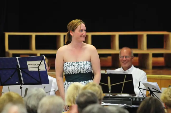 A woman stands beside seated musicians and music stands before an audience in an indoor performance space.
