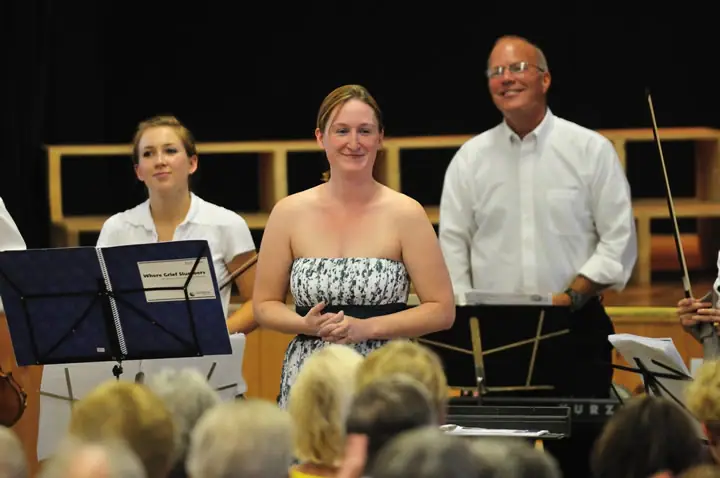 A singer and musicians stand onstage behind music stands as an audience watches.