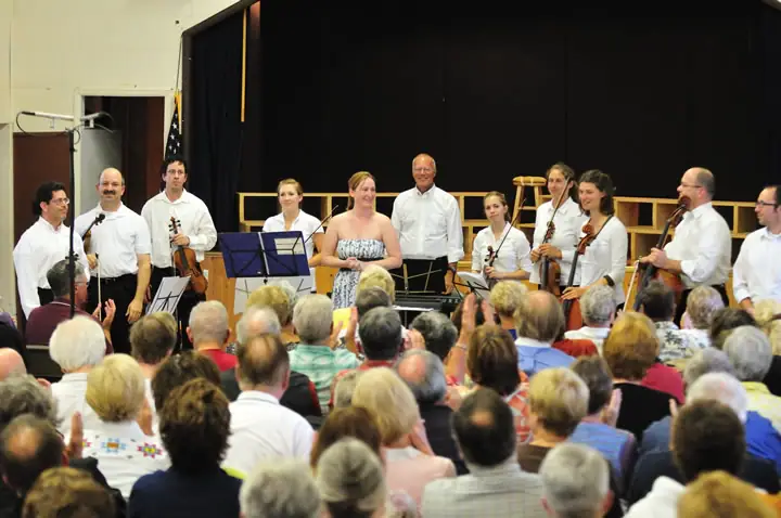 String musicians stand onstage before a seated audience in a crowded indoor concert hall.