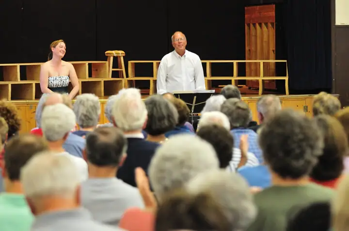Two people stand on a stage before a seated audience, with a wooden set and music stand behind them.