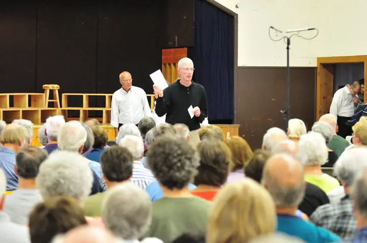 A man holds papers while addressing a seated audience in an auditorium.
