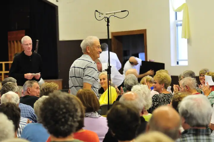 A man stands at a microphone before a seated audience in a crowded room.