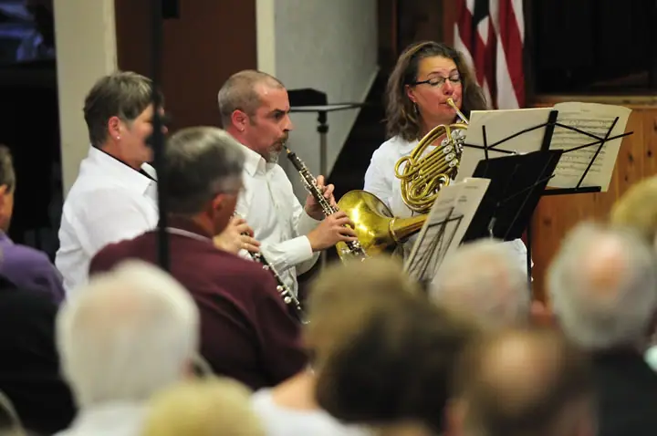 Musicians perform behind music stands while an audience watches in the foreground.
