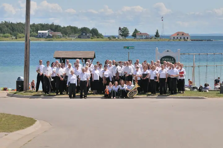 A large group of musicians poses beside a waterfront, with instruments, buildings, and a dock behind them.