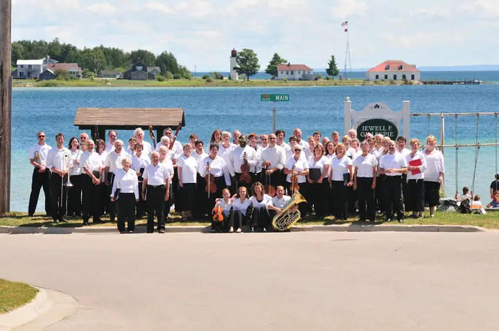 Large group of musicians posing beside a waterfront road with instruments, buildings, and a lighthouse behind them.