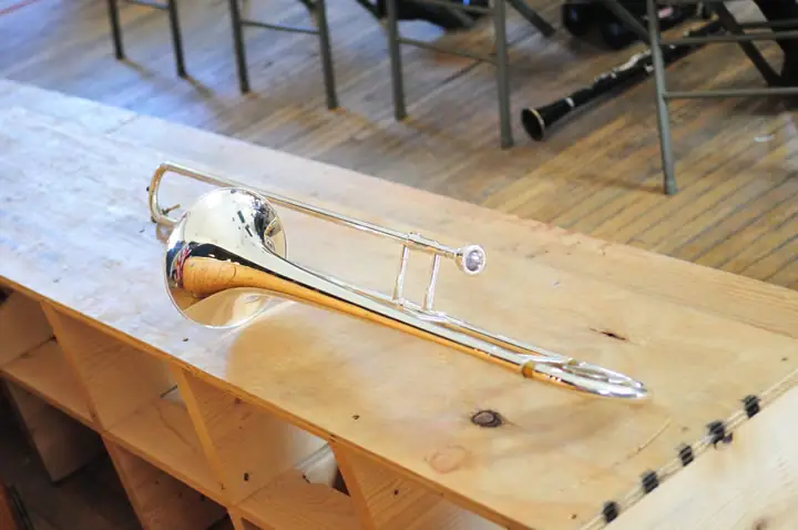 A silver trombone rests on a wooden bench in a room with folding chairs and another instrument.