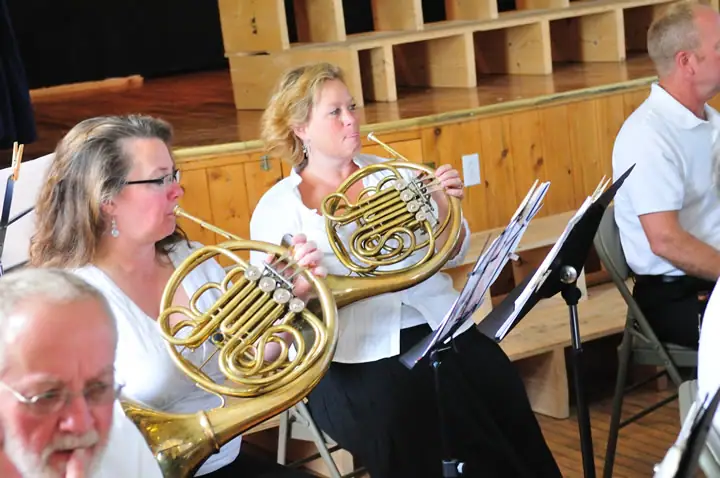 Two women play French horns alongside other musicians in a wood-paneled performance room.