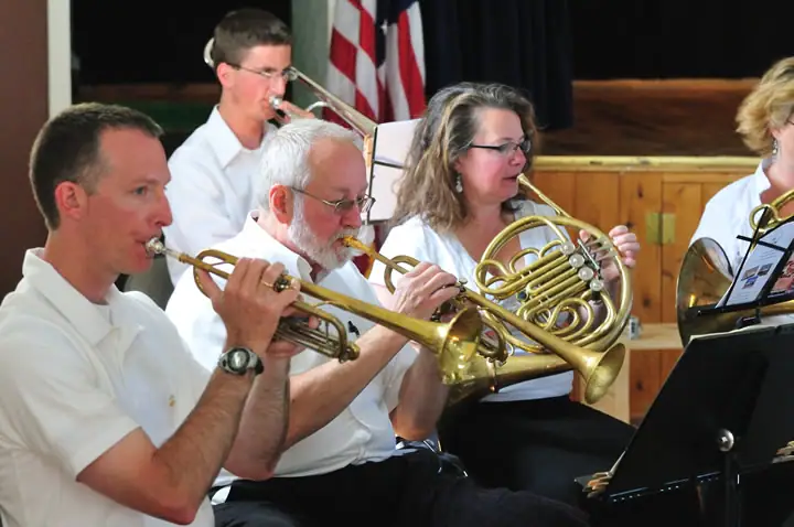 Five musicians play brass instruments on a stage, with music stands and flags behind them.