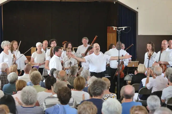 A conductor leads a string ensemble onstage before a seated audience.