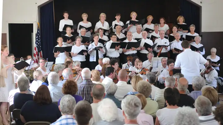 A choir and seated instrumentalists perform onstage before an audience, led by a conductor.