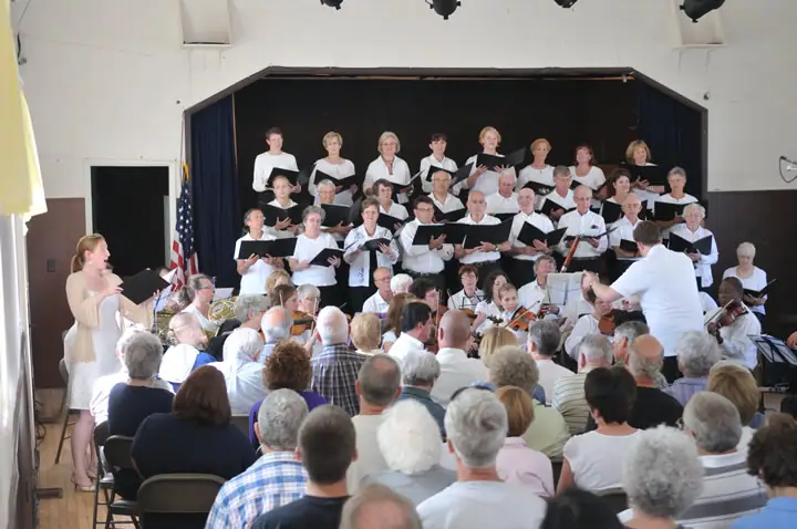 A choir performs before a seated audience, directed by a conductor in a white shirt.