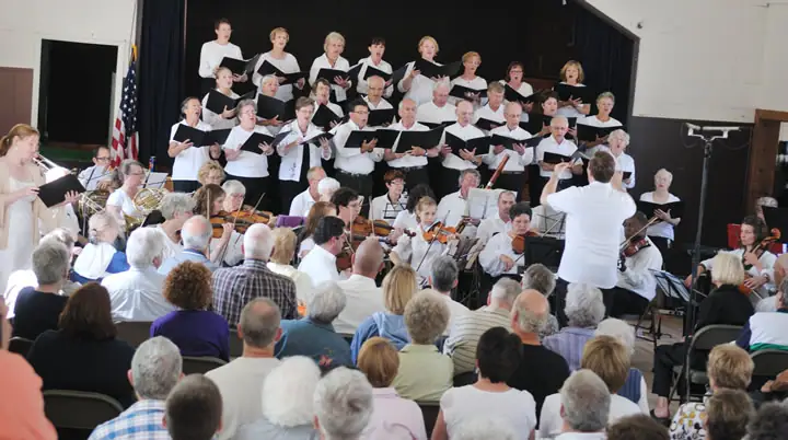 A choir and orchestra perform onstage before a seated audience, directed by a conductor.