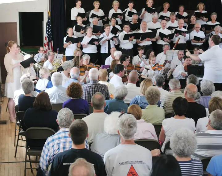 A choir and orchestra perform before a seated audience in an indoor hall.