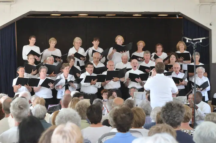 A choir performs on a small stage while a conductor leads from in front of an audience.