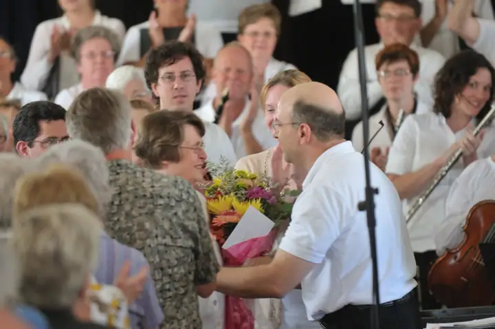 A man presents a bouquet to a woman while musicians and applauding people sit behind them.