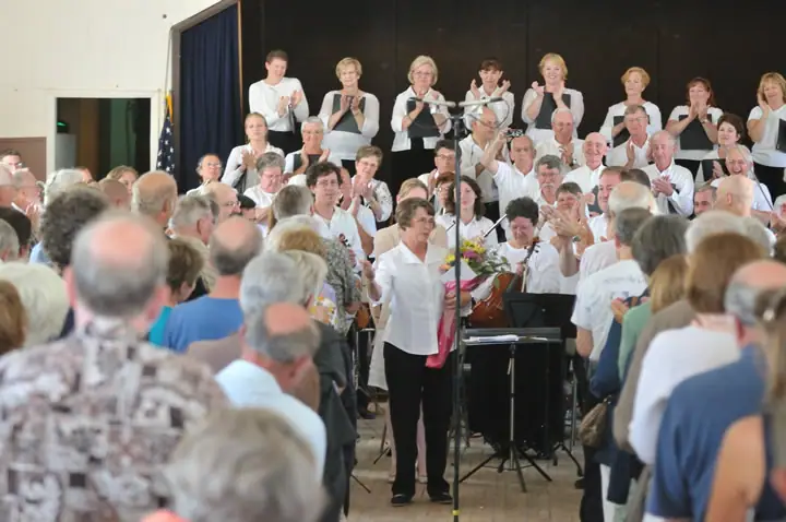 A crowd watches a large ensemble applauding around a woman holding flowers and standing beside a microphone.