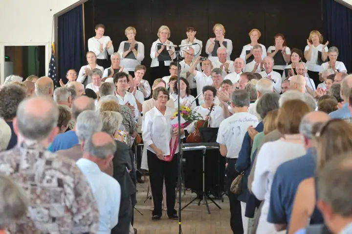 A choir and audience gather indoors as a woman holding flowers stands at a microphone.