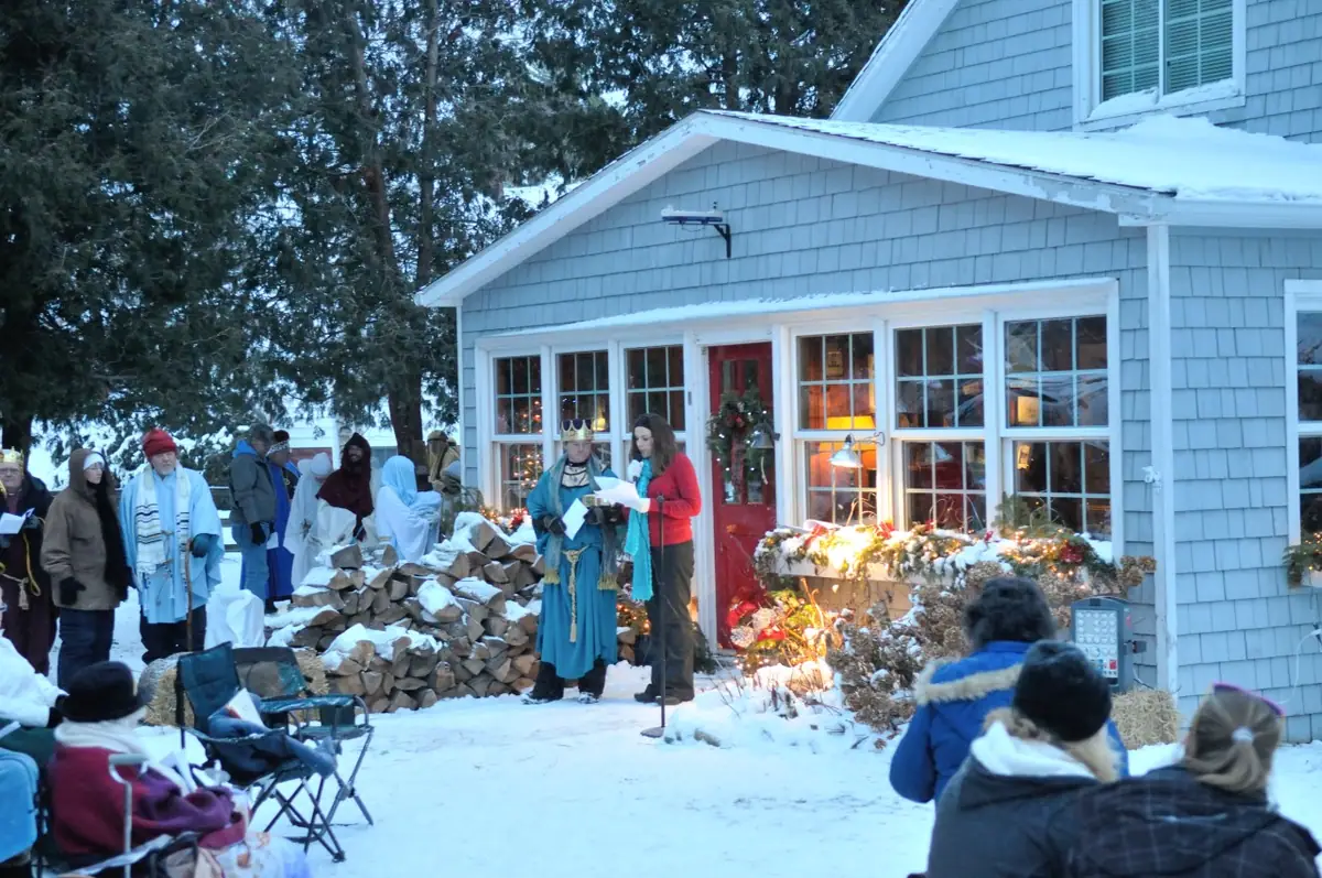 A costumed group gathers outside a shingled house in the snow while seated spectators watch.